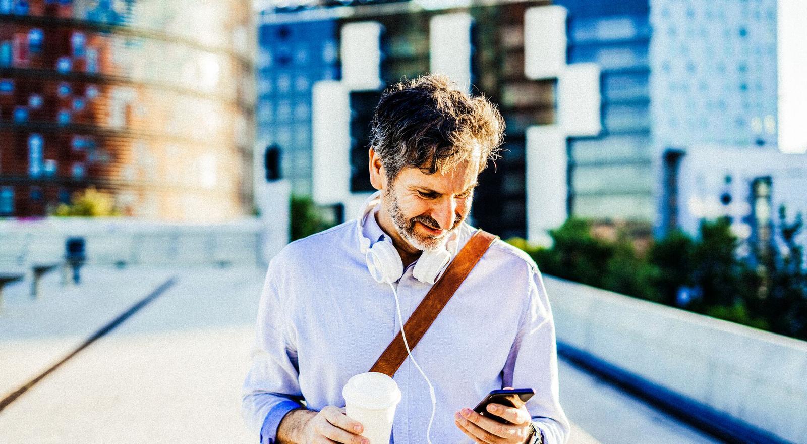 Mid adult man holding coffee cup looking at mobile phone with headphones around neck Mid adult man holding coffee cup looking at mobile phone with headphones around neck