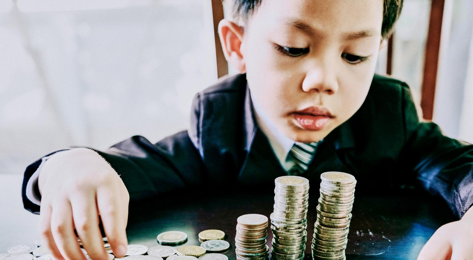 A young boy in a suit counts and stacks coins on a table