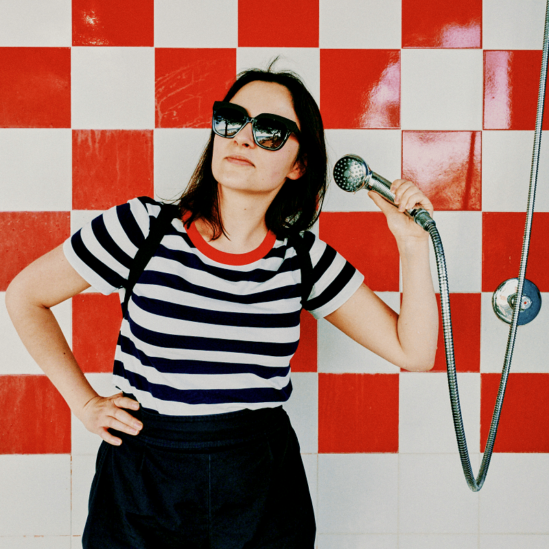 A woman with sunglasses pretends to pick up the phone by using the shower head in a retro shower