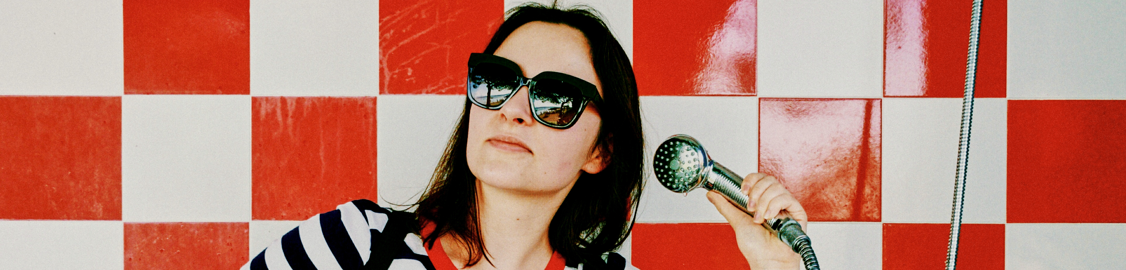 Hero A woman with sunglasses pretends to pick up the phone by using the shower head in a retro shower crop